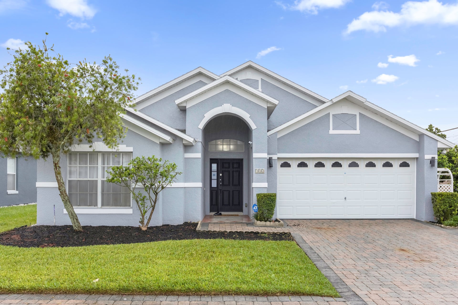 Exterior view of a light blue single-story house with a white garage door, paved driveway, and a landscaped front yard.