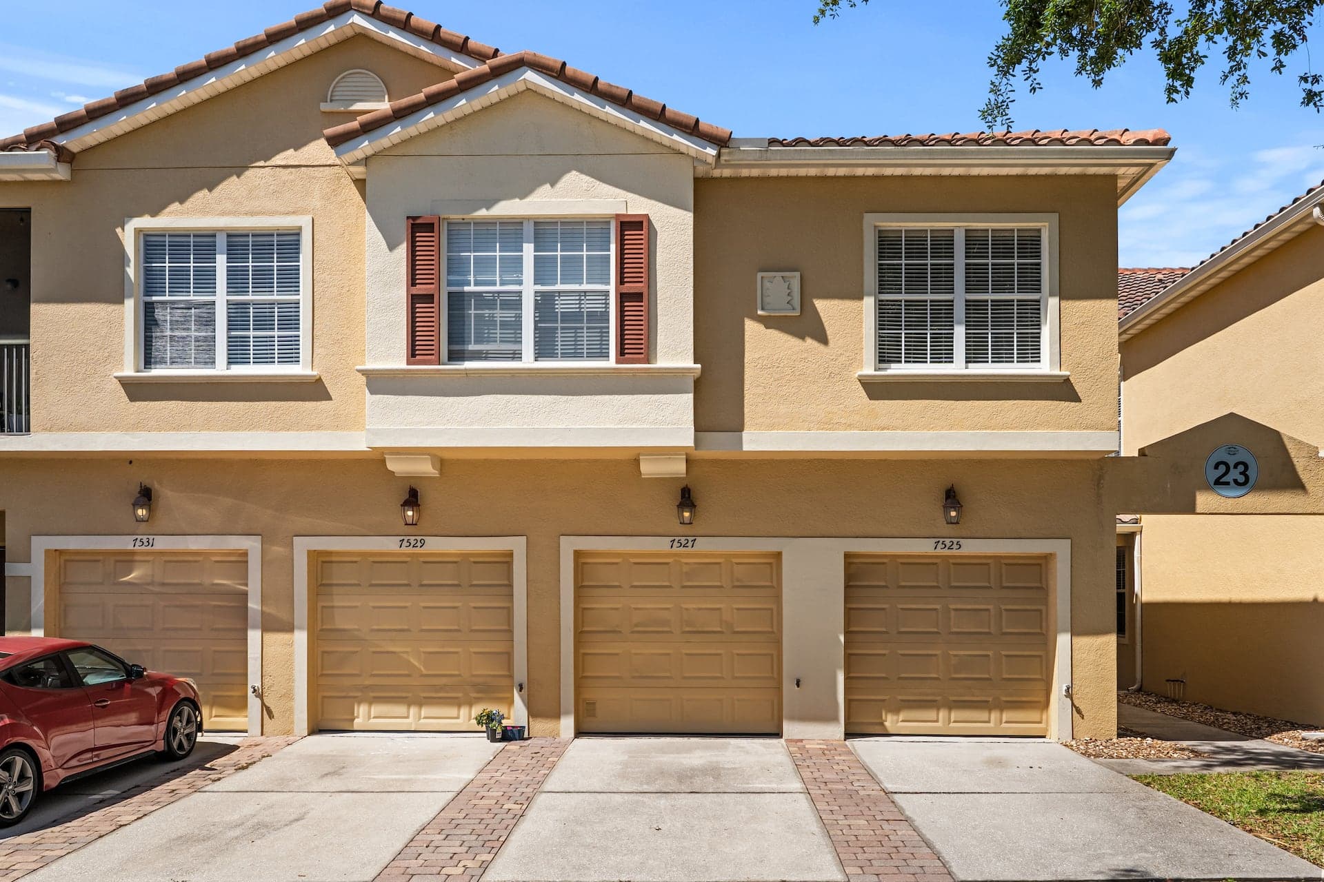 Front exterior of a multi-unit townhouse building featuring beige stucco, a tiled roof, four individual garage doors with paved driveways, and a building number sign.