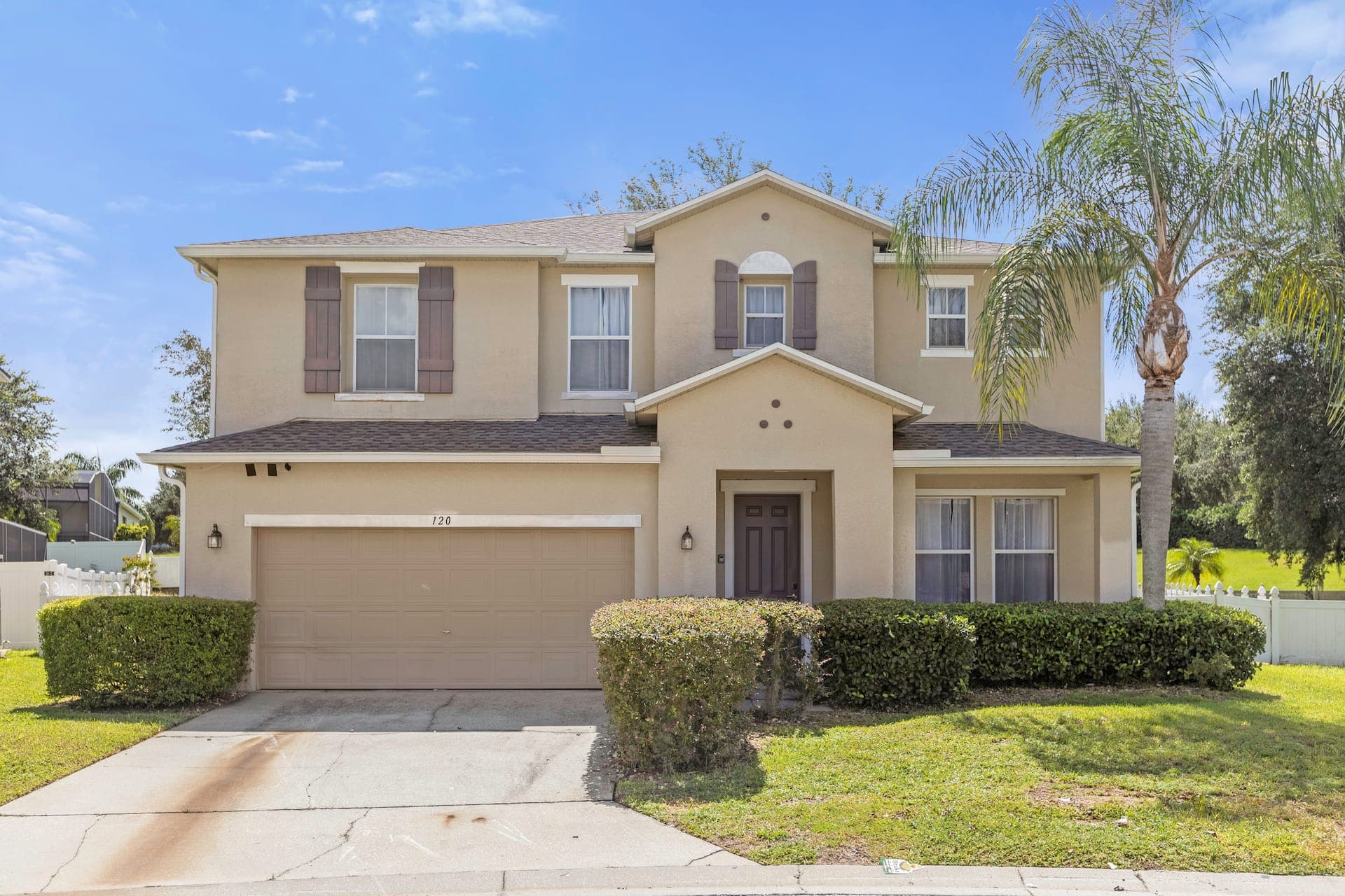 Exterior view of a two-story beige house featuring a brown garage door, manicured hedges, and a palm tree.