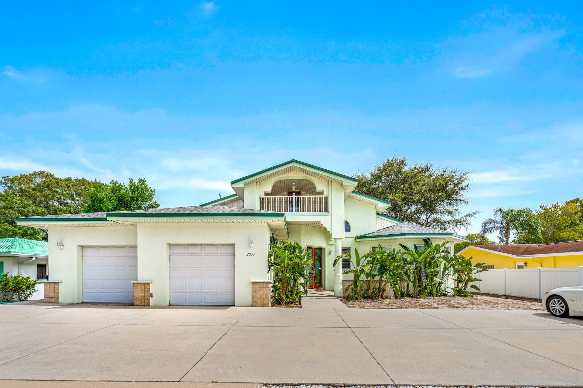 Exterior view of a large, modern two-story vacation home with beige stucco, brown trim, a paved driveway, and landscaped yard under a bright sky.