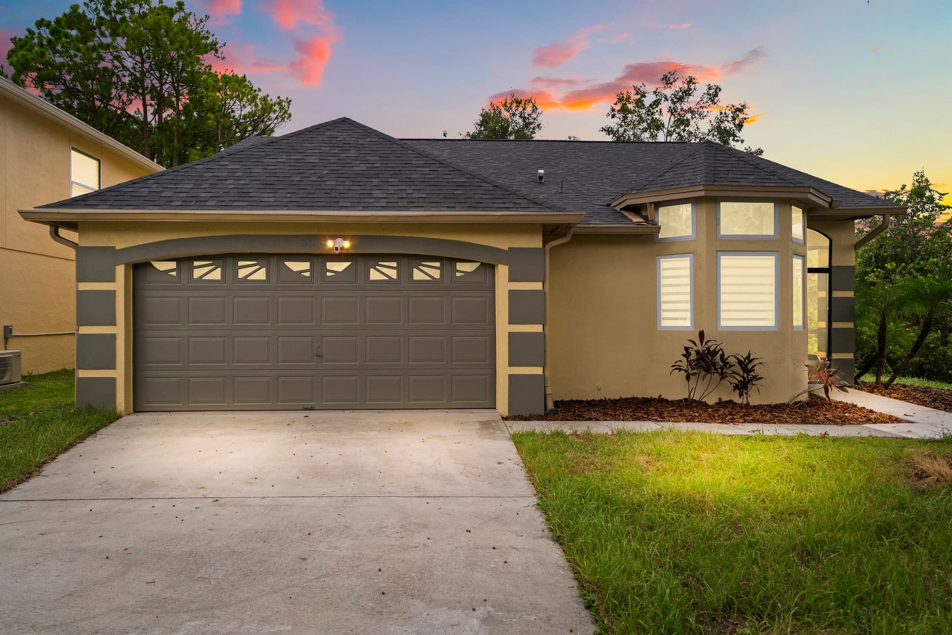 Exterior view of a tan single-story house with grey trim, a bay window, and a brown garage door at sunset.