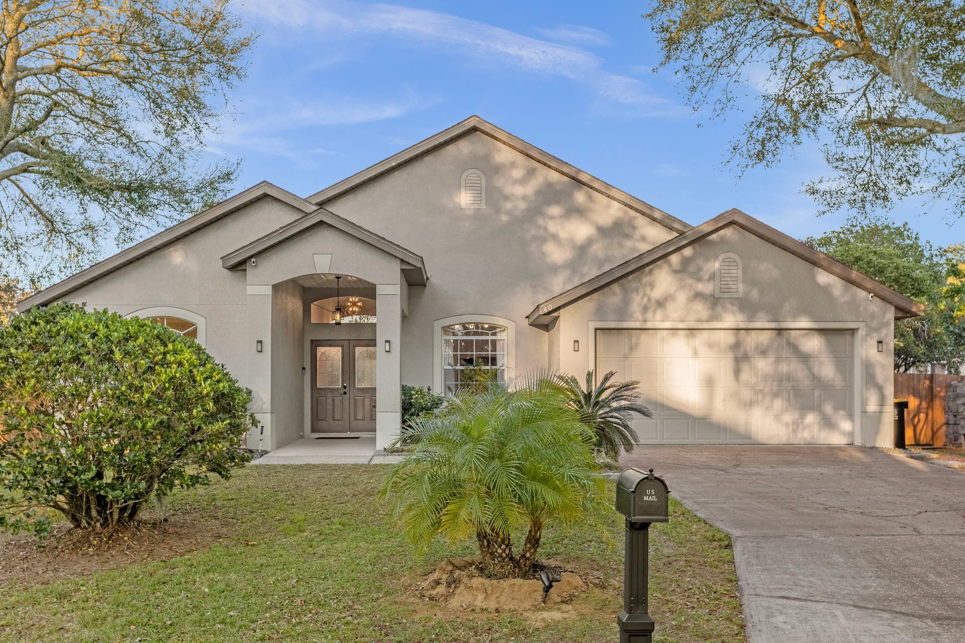 Front view of a single-story beige house with a two-car garage, double front doors, and a small palm tree on the front lawn during the day.