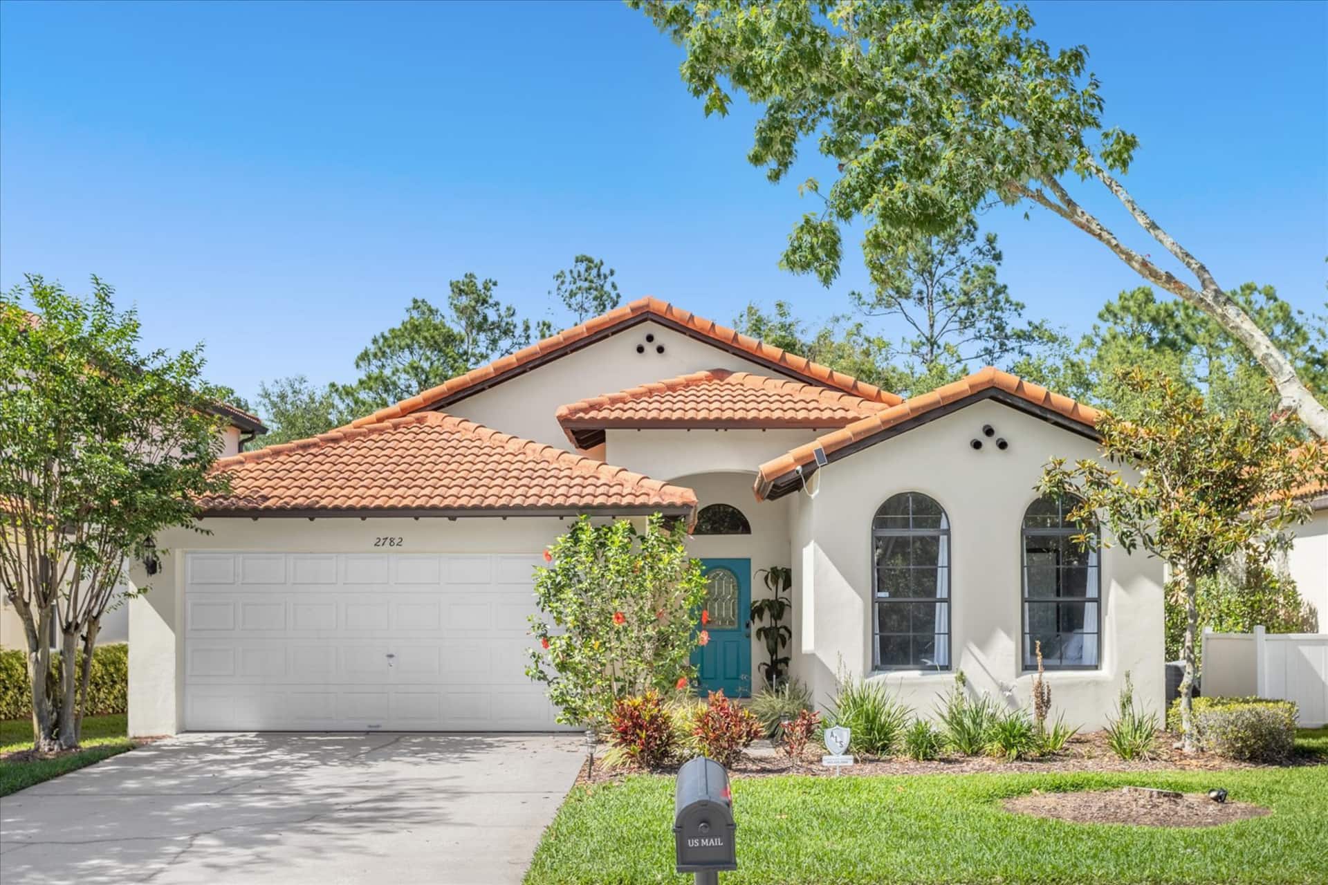 Front exterior of a single-story, beige stucco home featuring a terracotta tile roof, arched windows, a bright teal front door, a two-car garage, and a landscaped front yard.