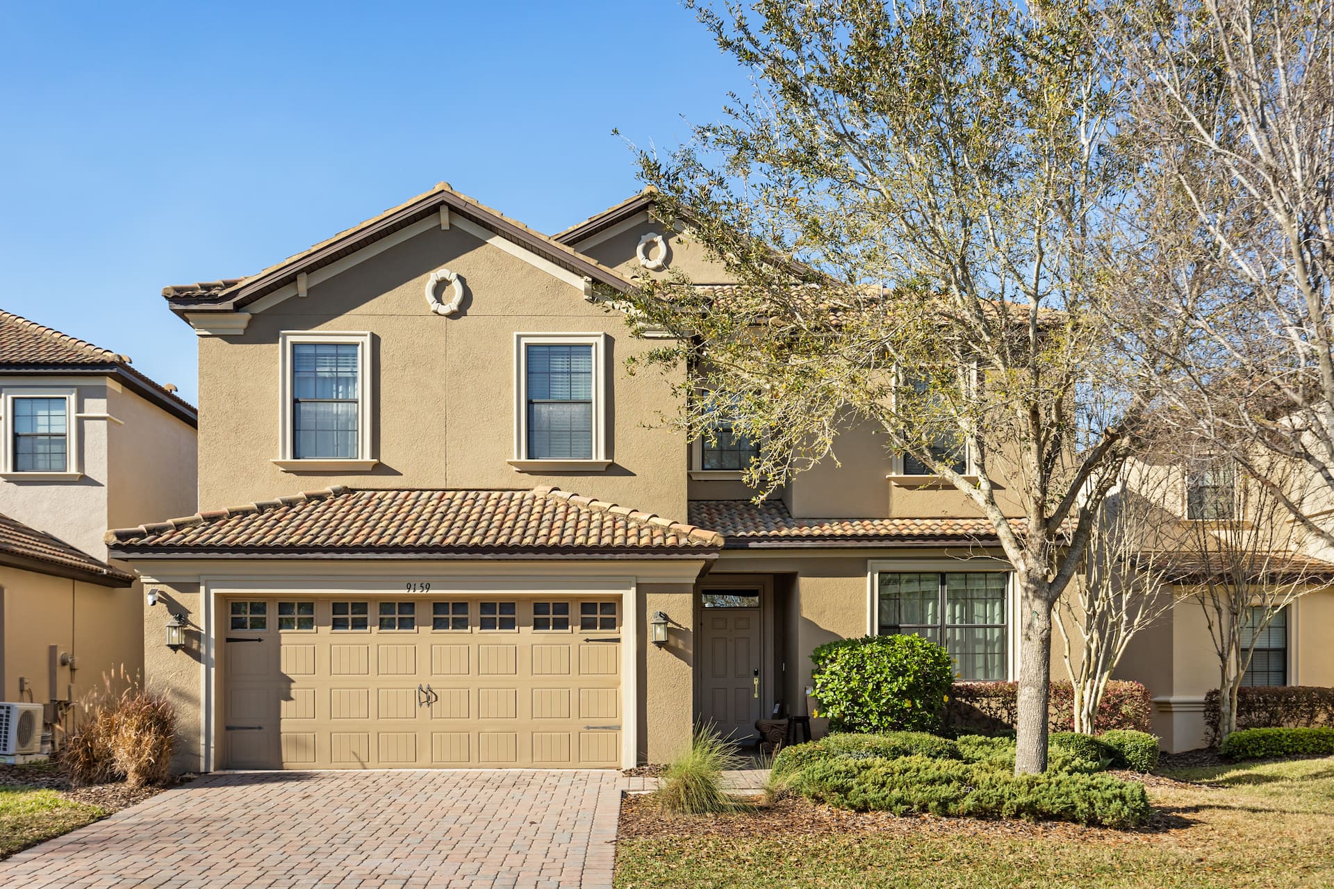 Daytime front exterior of a two-story beige stucco home featuring a tiled roof, a two-car garage, and a mature tree in the landscaped front yard.