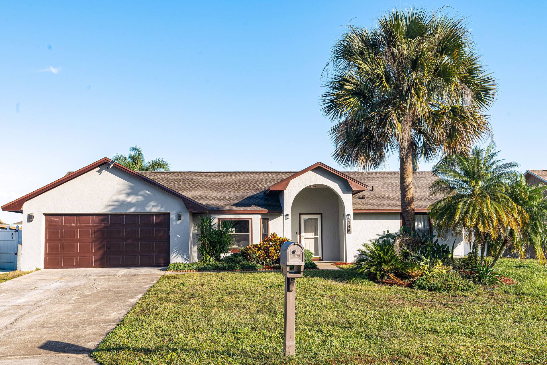 Front exterior of a single-story house featuring a brown garage door, an arched entryway, and a large palm tree in the front yard under a clear blue sky.